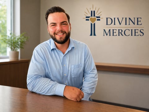 Man in light blue shirt smiling at desk with Divine Mercies logo on wall behind him